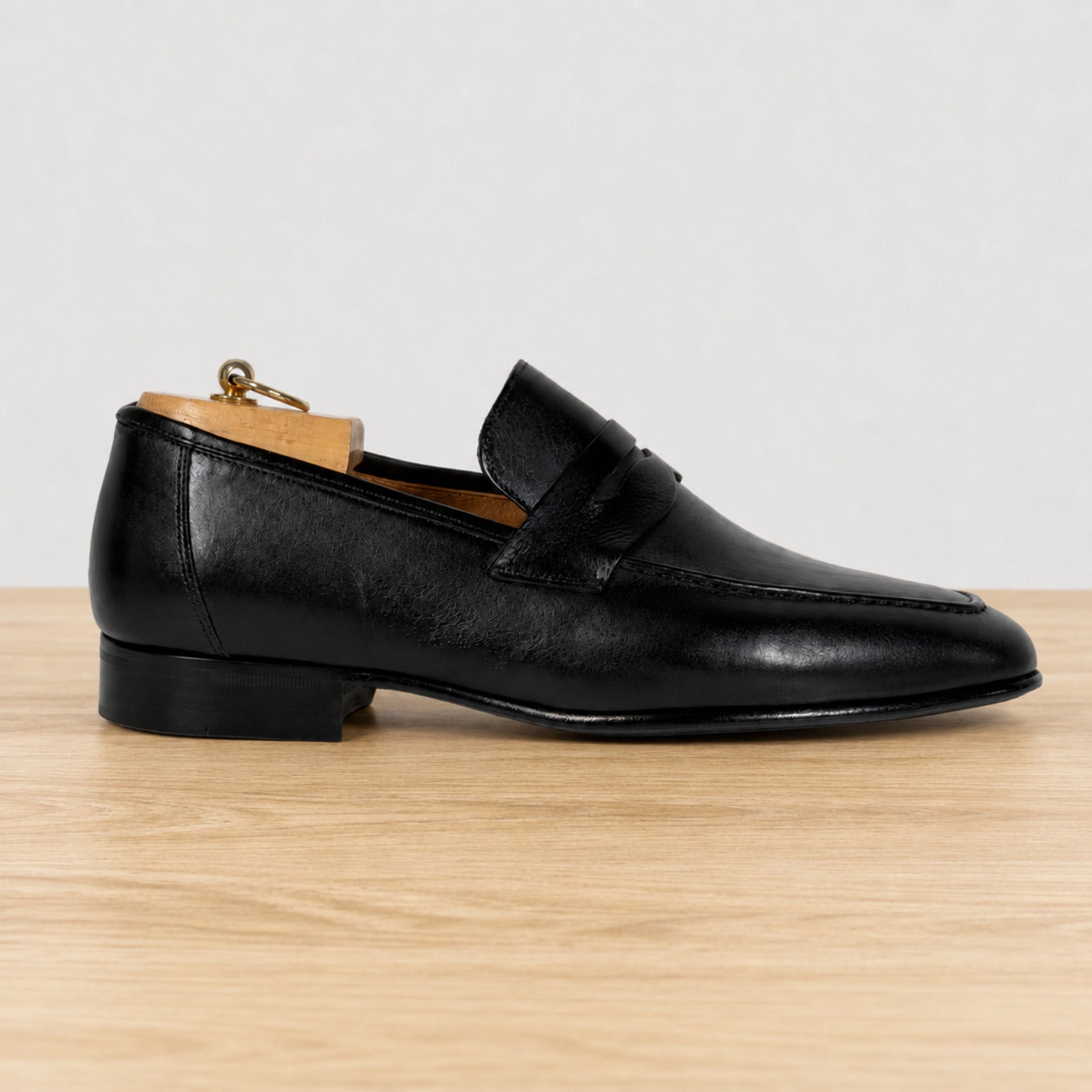 A black leather penny loafer shoe with a low stacked heel, displayed on a light wood surface against a neutral light gray background.