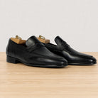 A pair of black leather penny loafers with a low block heel, displayed on a light wood surface against a neutral off-white background.