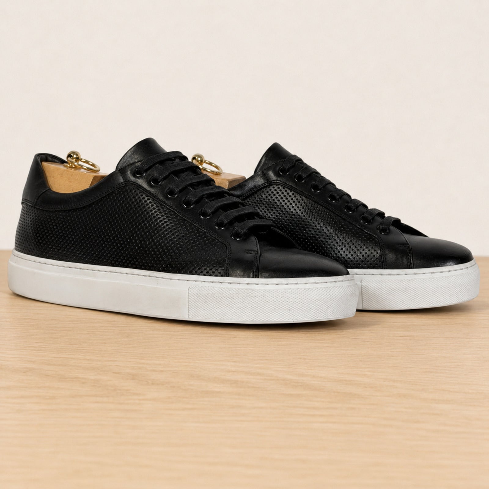 A pair of black leather low-top sneakers with perforated detailing, black laces, and white rubber soles, displayed on a light wood surface against a warm beige background.