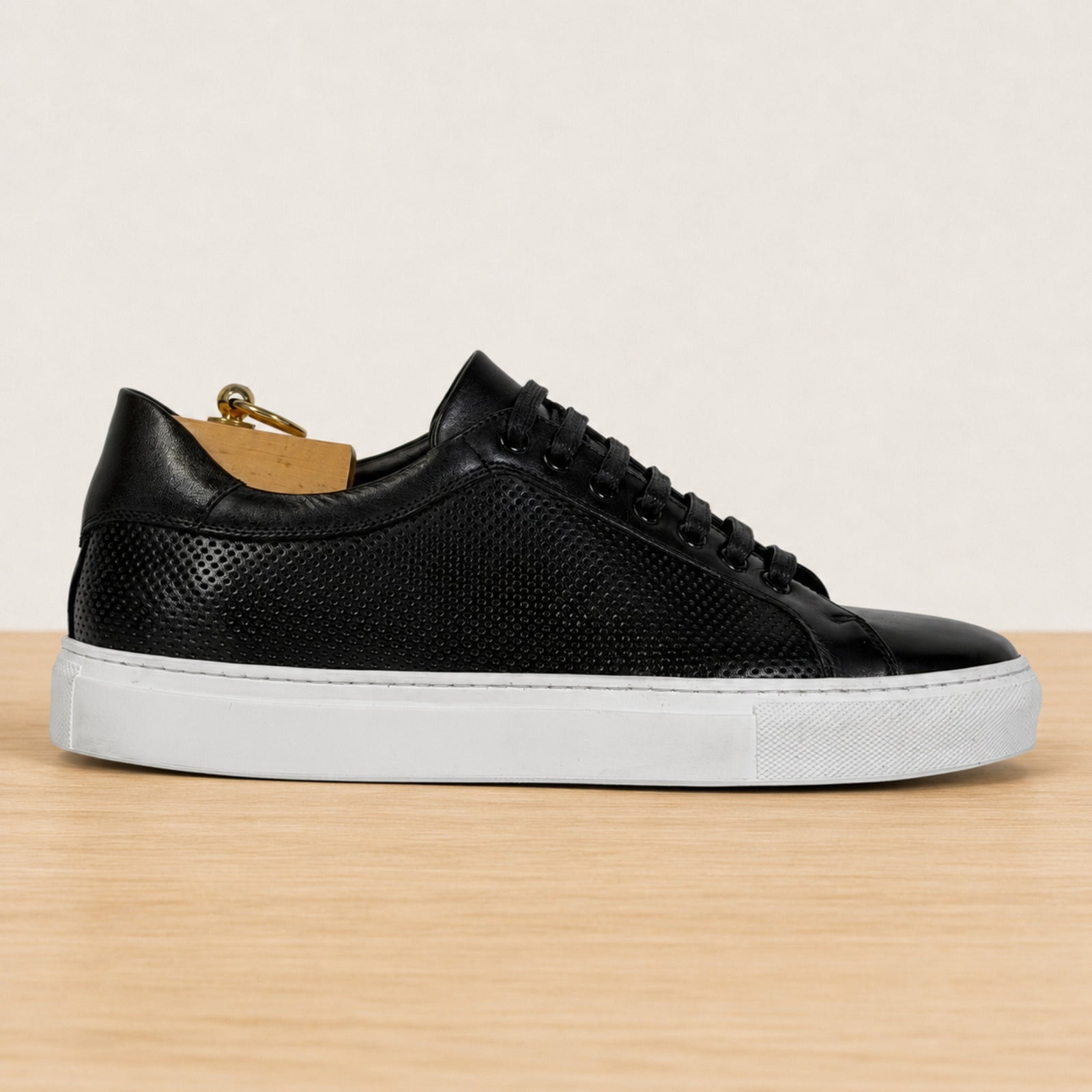 A black leather low-top sneaker with a textured perforated upper, black laces, and a thick white rubber sole, displayed on a light wood surface against a neutral beige background.