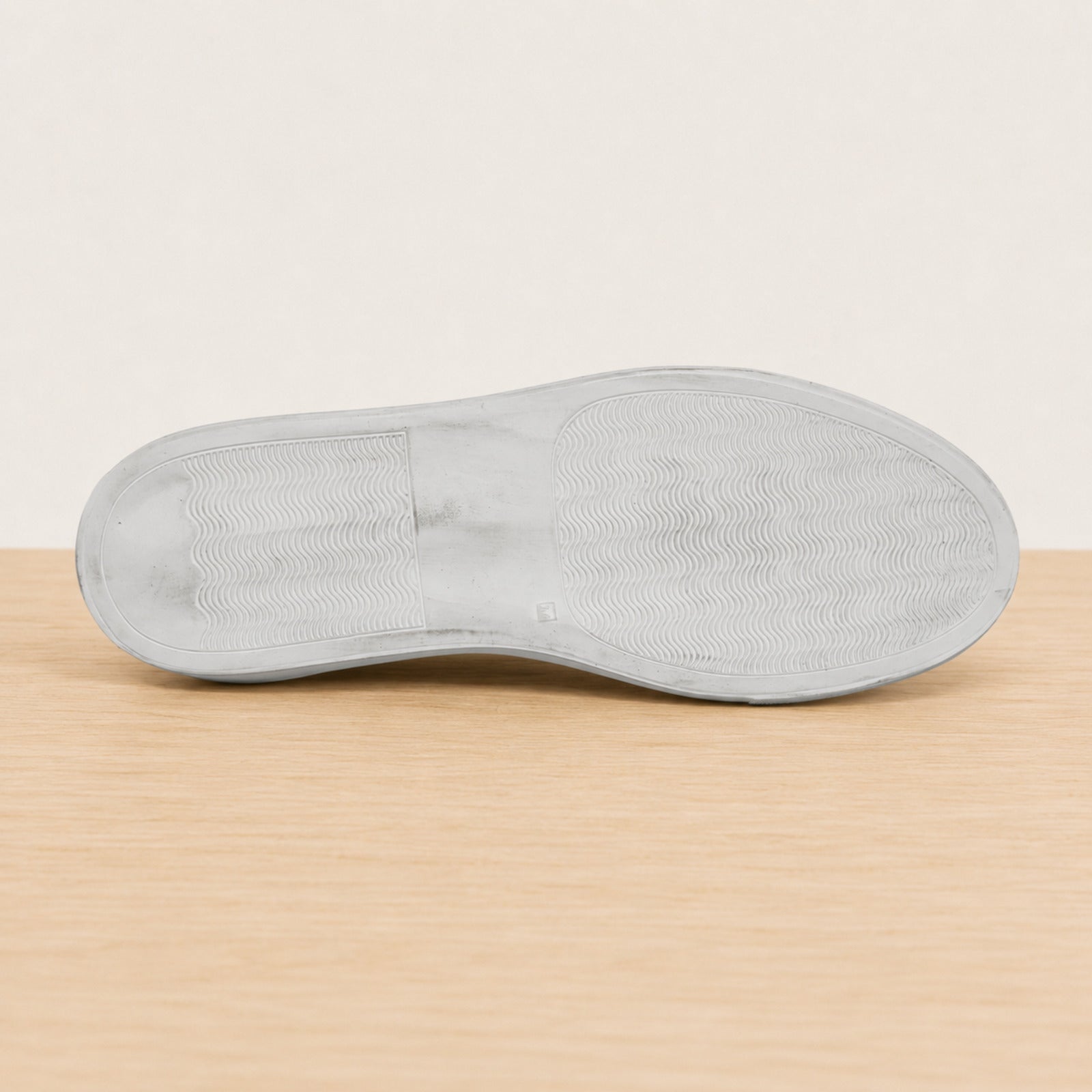 The outsole of a light gray shoe shown from a top-down view, resting on a light wood surface against a plain off-white background.