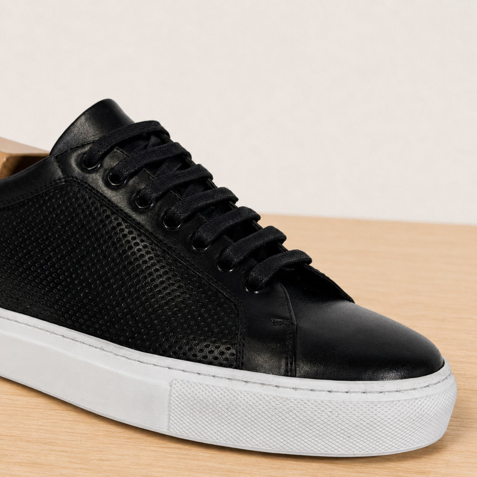 A black leather and perforated sneaker with black laces and a thick white sole, shown on a light wood surface against a neutral beige background.