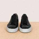 A pair of black leather low-top sneakers with perforated side panels and light gray rubber soles, displayed on a light wood surface against a beige background.