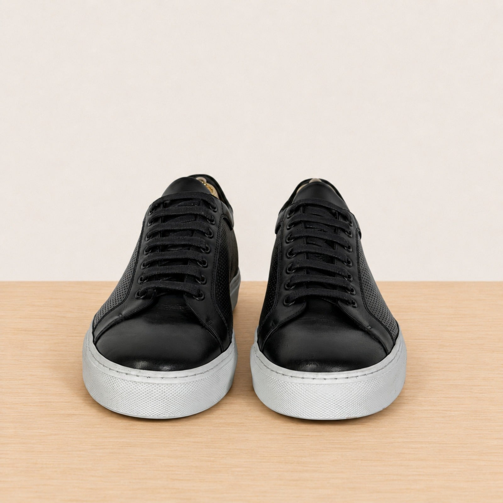 A pair of black leather low-top sneakers with perforated side panels and light gray rubber soles, displayed on a light wood surface against a beige background.