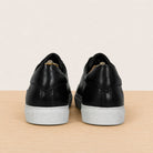 A pair of black leather low-top sneakers shown from the back, featuring textured panels, black laces, and light gray rubber soles, set against a neutral beige background and light wood floor.