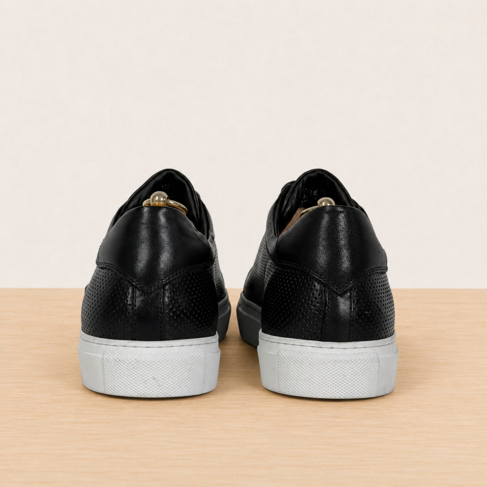 A pair of black leather low-top sneakers shown from the back, featuring textured panels, black laces, and light gray rubber soles, set against a neutral beige background and light wood floor.