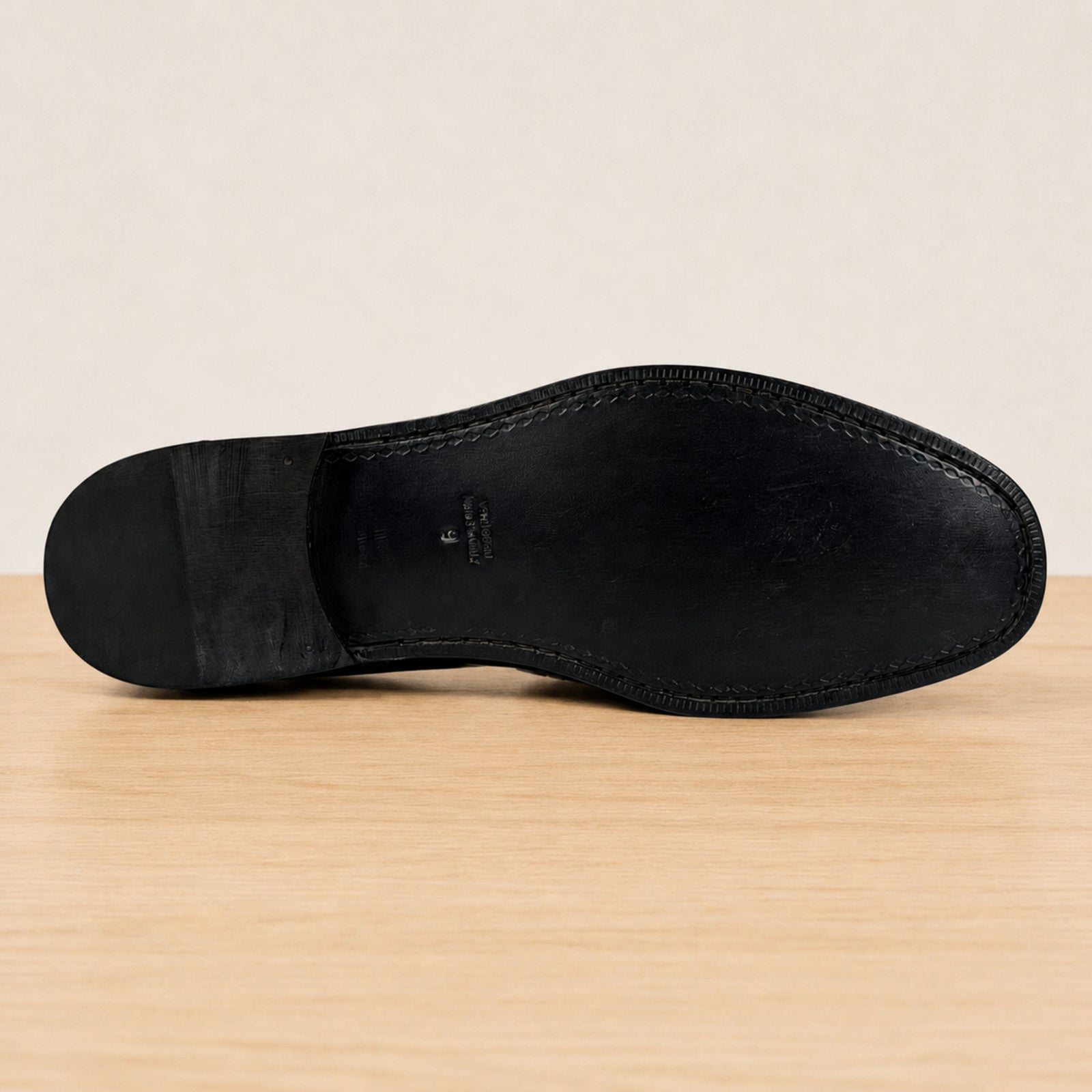The black leather underside of a dress shoe sole, showing the heel, toe, and stitched edge, resting on a light wood surface against a neutral beige background.