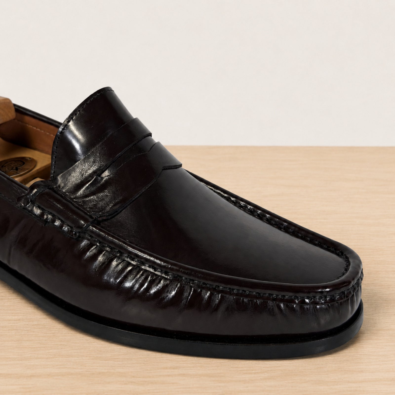 A close-up of a glossy burgundy leather penny loafer resting on a wooden surface against a light gray background.
