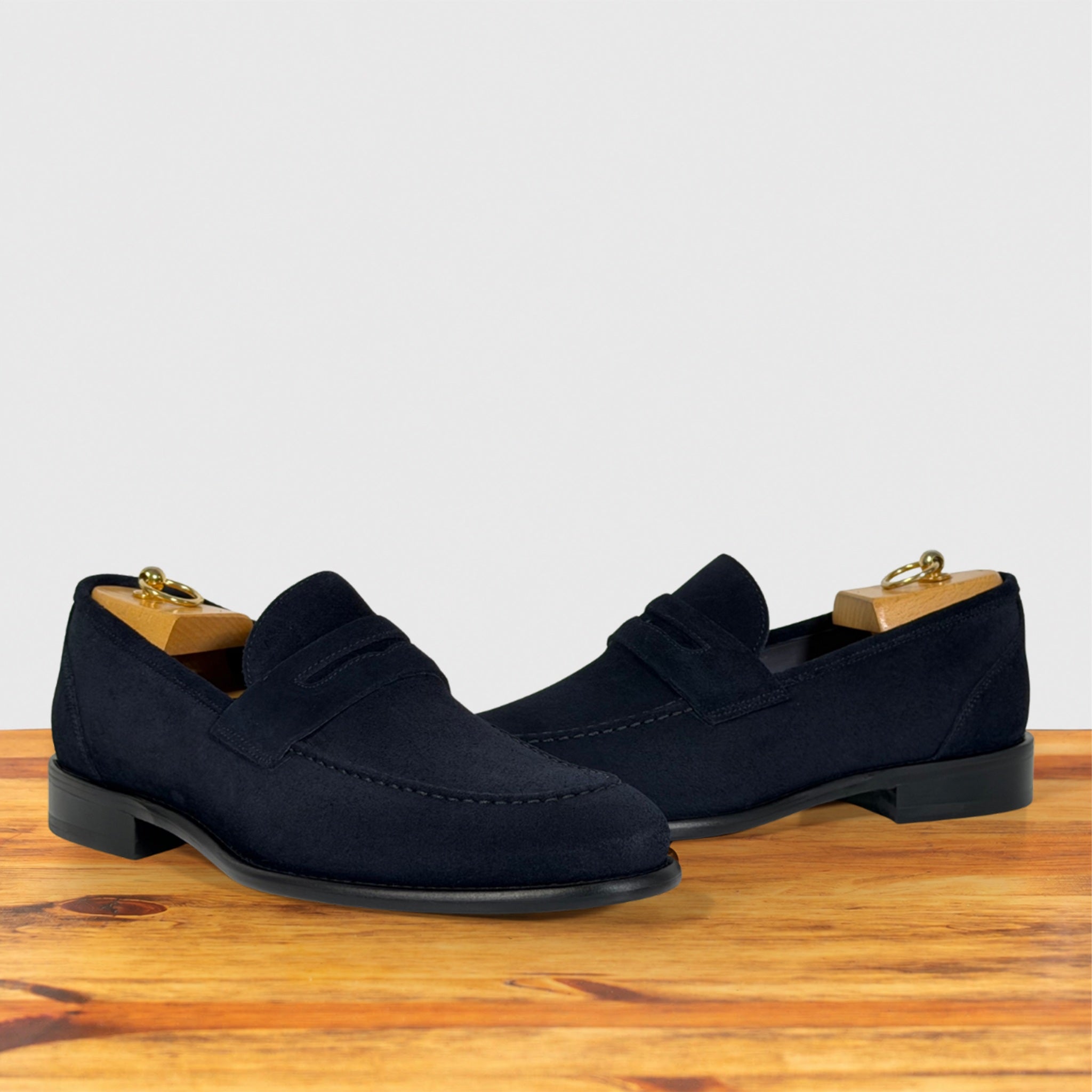 A pair of dark navy blue suede penny loafers displayed with wooden shoe trees, set against a light gray background and resting on a wooden surface.