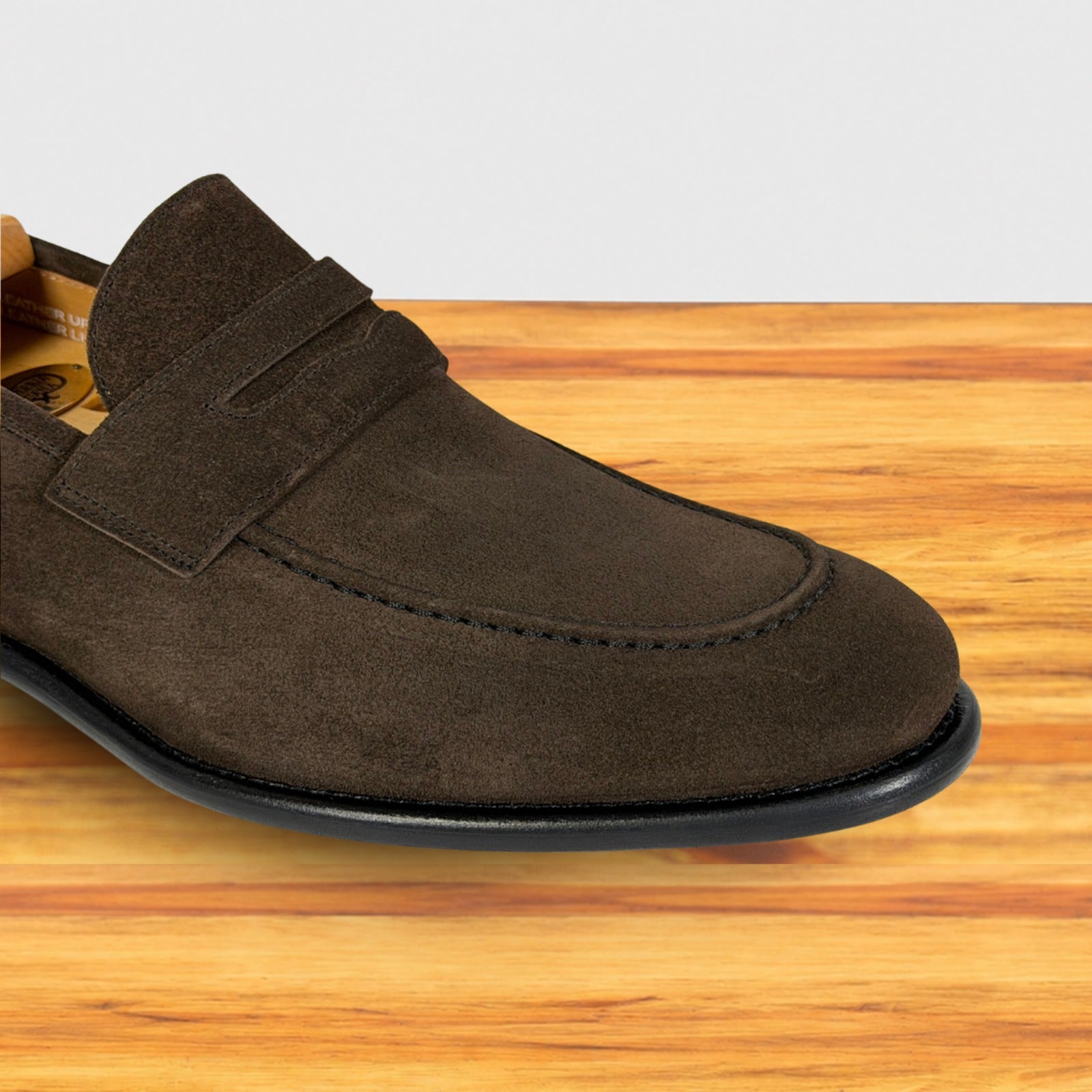 A dusty brown suede penny loafer shown from a side profile view, resting on a light brown wooden surface against a white background.