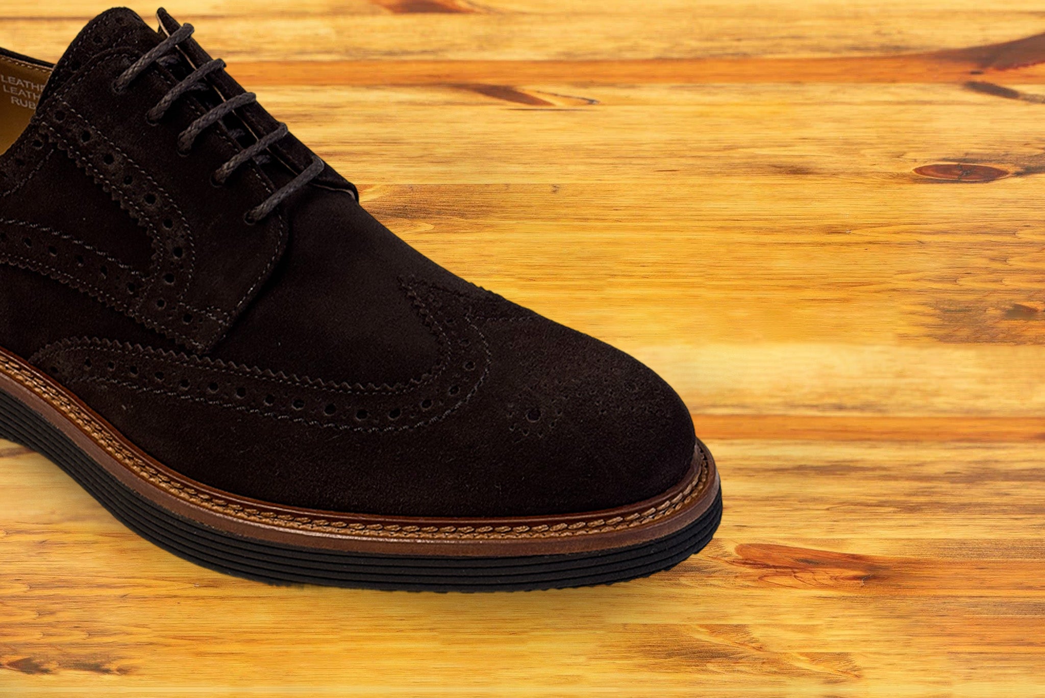 A brown suede wingtip shoe with a rubber sole, displayed on a wooden surface.