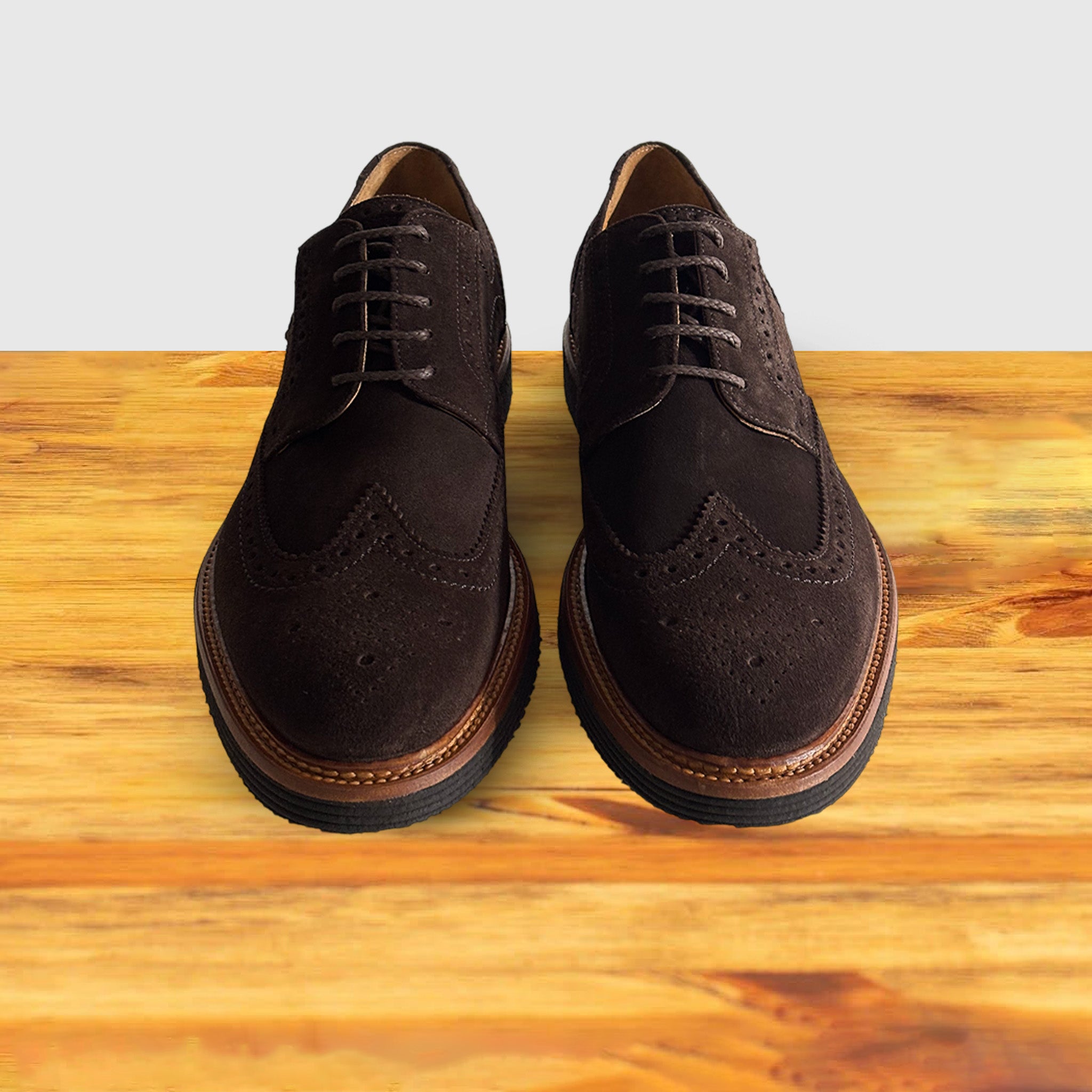 A Pair of brown suede wingtip shoe with a rubber sole, displayed on a wooden surface.