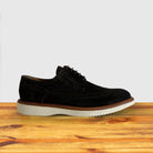 A Olive Green suede wingtip derby shoe with a rubber sole, displayed on a wooden surface.