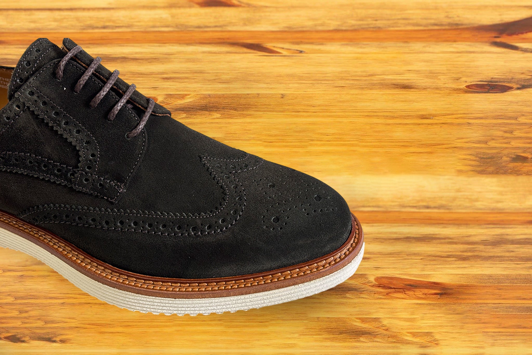A Olive Green suede wingtip derby shoe, displayed on a wooden surface.