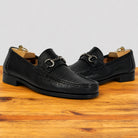 A pair of black leather penny loafers with a horsebit detail, displayed on a wooden surface against a light gray background.