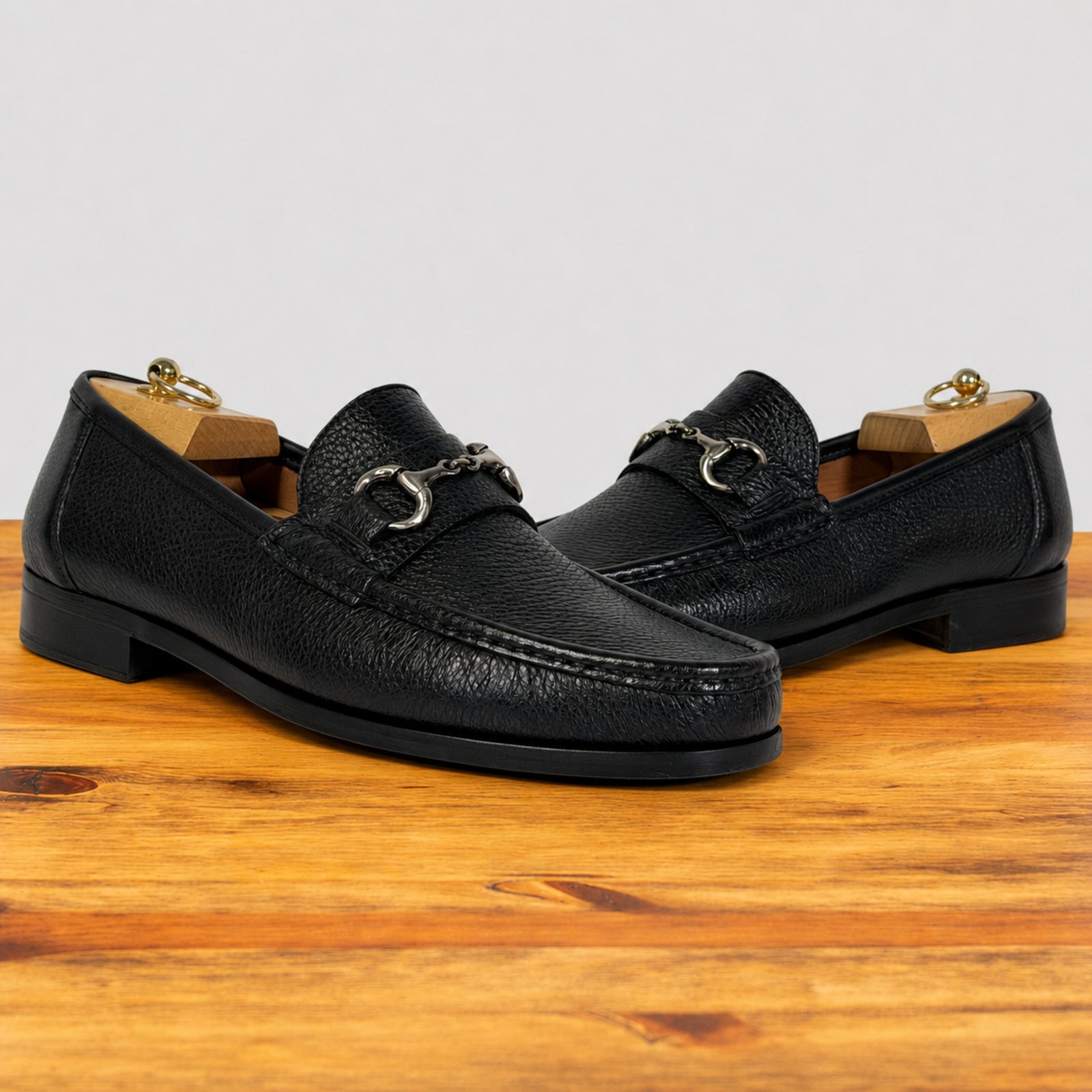 A pair of black leather penny loafers with a horsebit detail, displayed on a wooden surface against a light gray background.