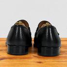 A pair of black leather penny loafers shown from the back, resting on a wooden surface against a light gray background.