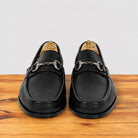 A pair of black leather penny loafers with silver horsebit hardware, displayed on wooden stands on a wooden surface against a light gray background.