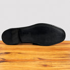 The black rubber outsole of a dress shoe, showing the Vastel logo embossed in the center, resting on a wooden surface against a light gray background.