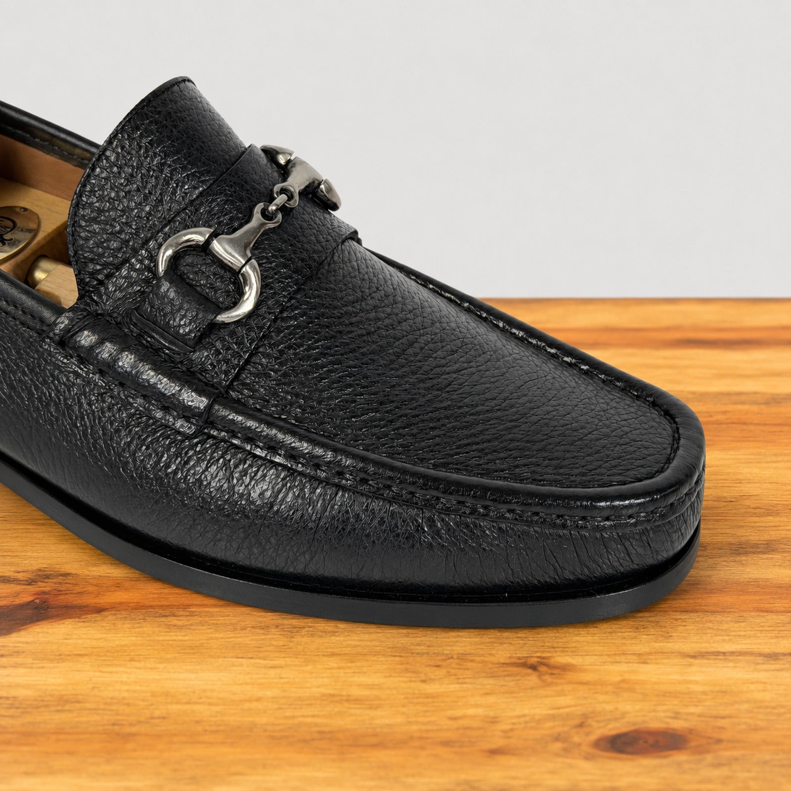 A close-up of a black pebbled leather loafer with a silver horsebit detail, resting on a wooden surface against a light gray background.