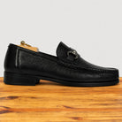 A single black pebbled leather loafer with a silver-tone horsebit detail, displayed on a wooden surface against a light gray background.