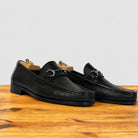 A pair of black leather loafers with a textured grain finish and a silver-tone horsebit detail on the vamp, displayed on a wooden surface against a light gray background.