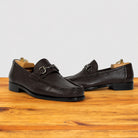 A pair of dark brown pebbled leather penny loafers with horsebit detailing, displayed on a wooden surface against a light gray background.