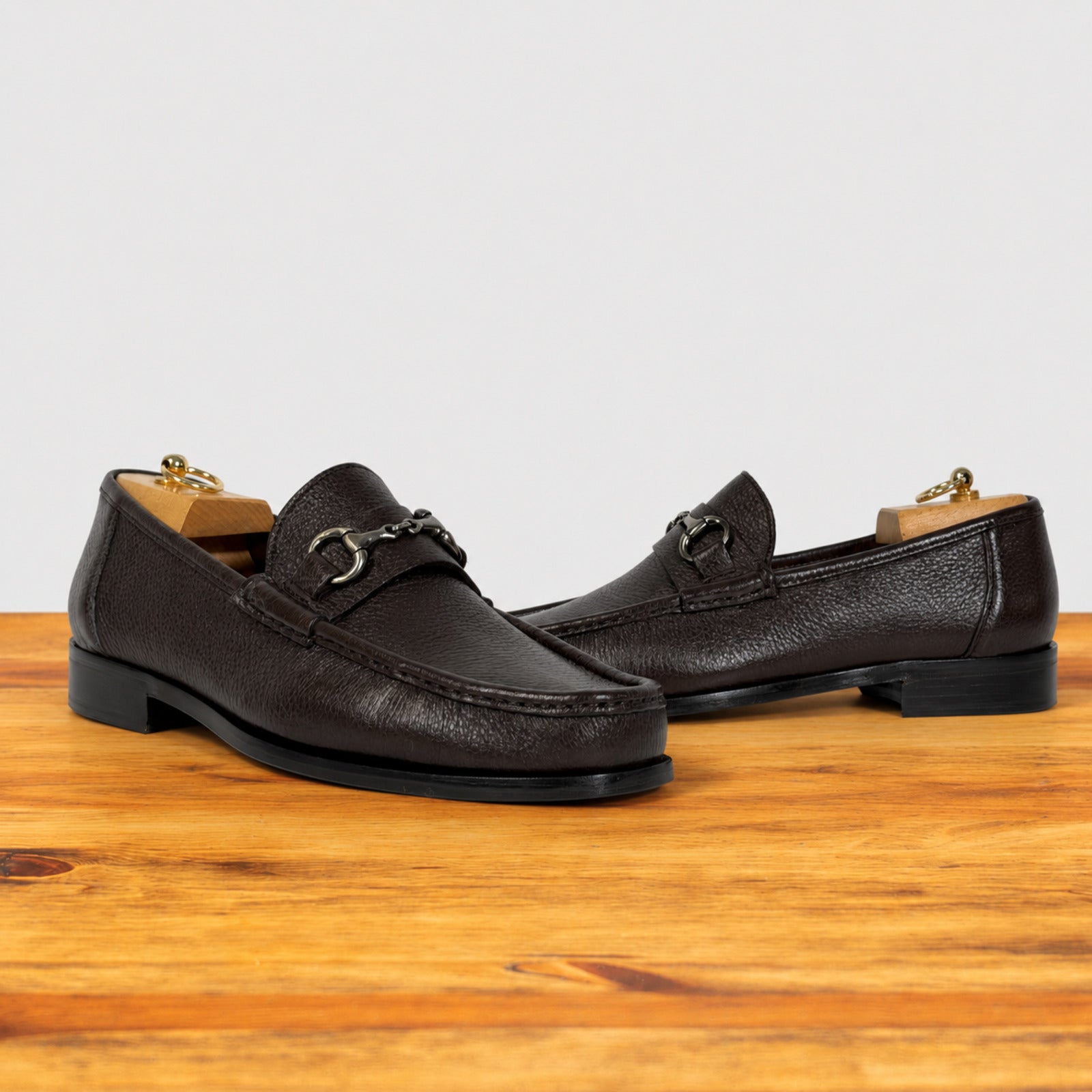 A pair of dark brown pebbled leather penny loafers with horsebit detailing, displayed on a wooden surface against a light gray background.