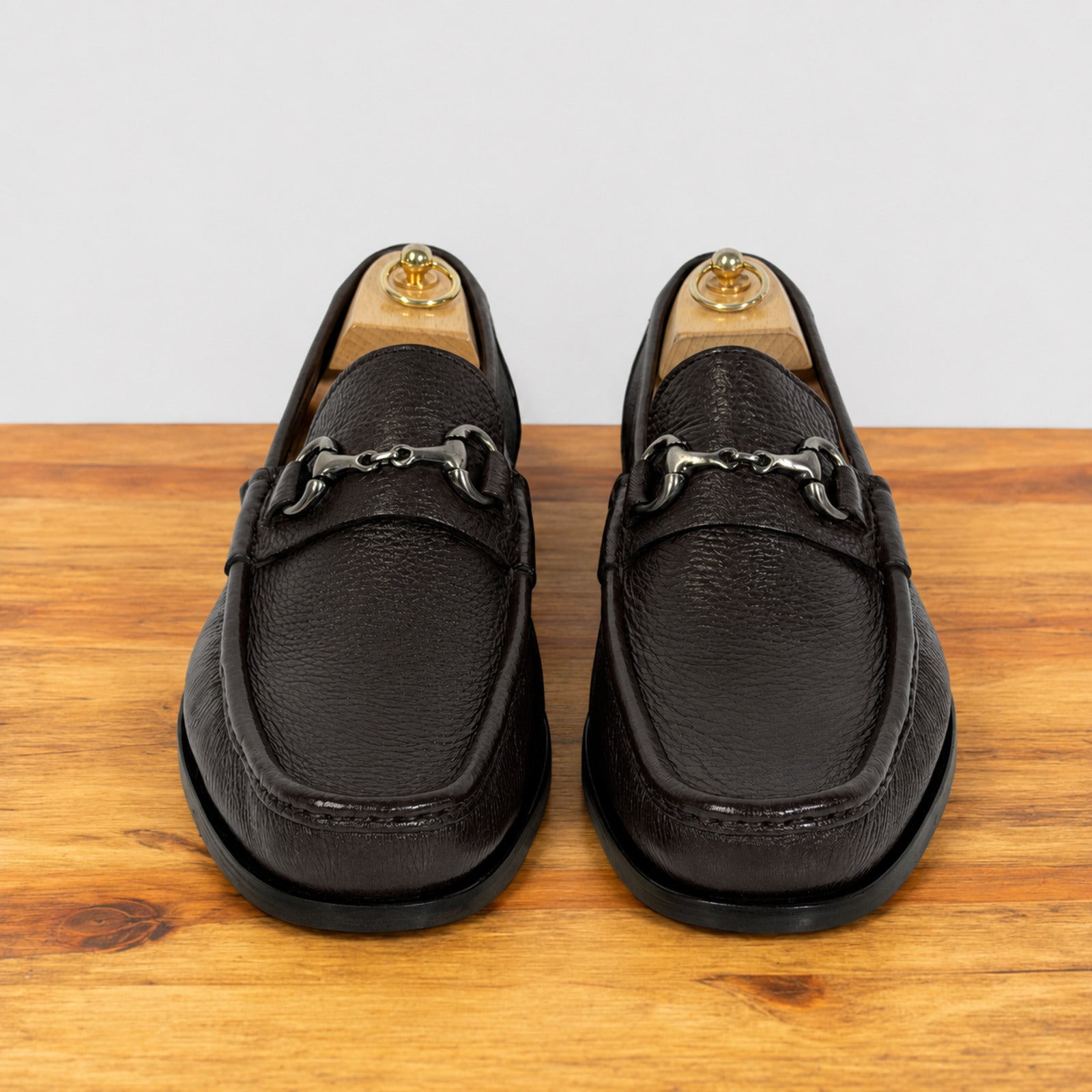 A pair of dark brown leather penny loafers with a horsebit detail, displayed on a wooden surface against a light gray background.