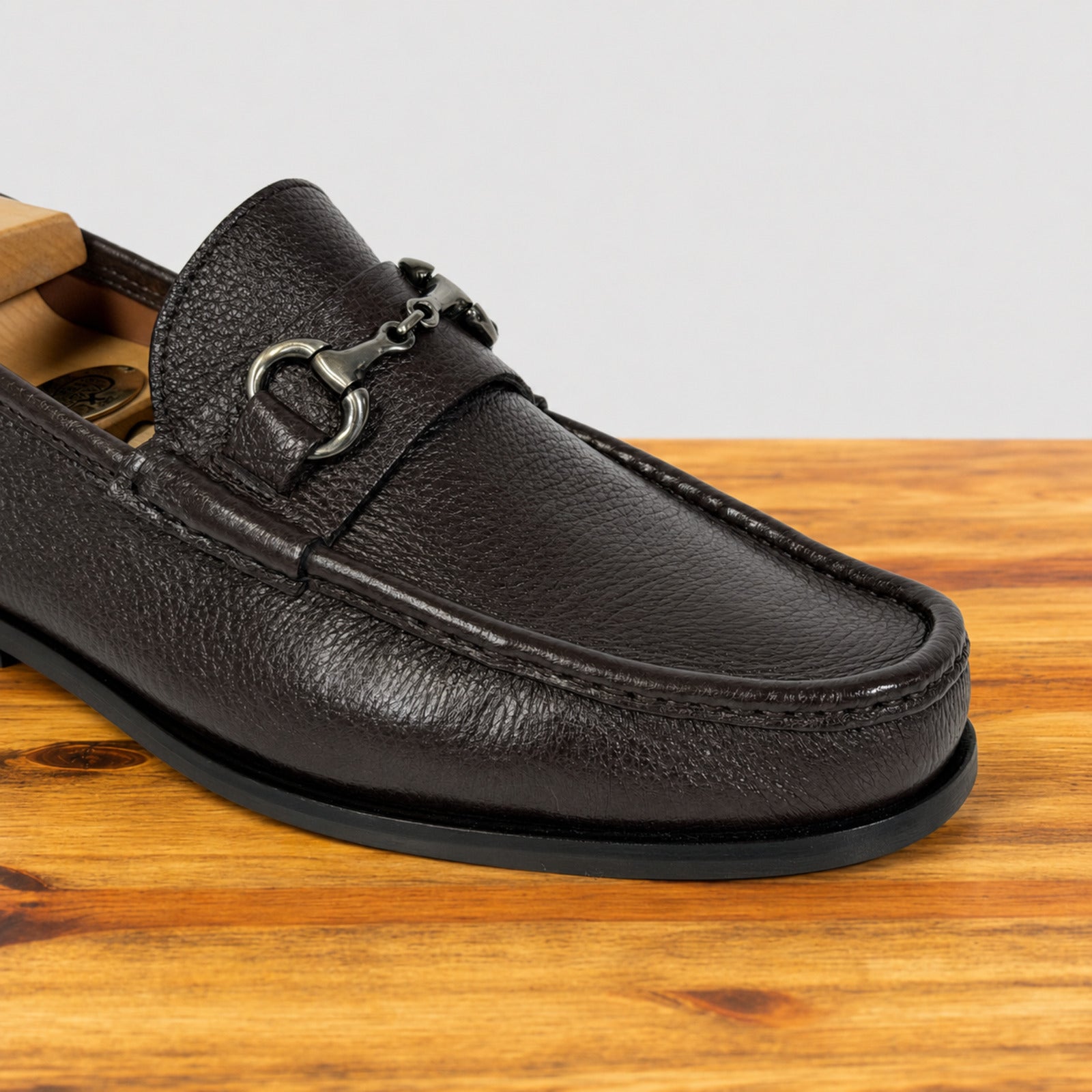 A close-up of a dark brown pebbled leather loafer with a silver-tone horsebit detail, resting on a wooden surface against a light gray background.