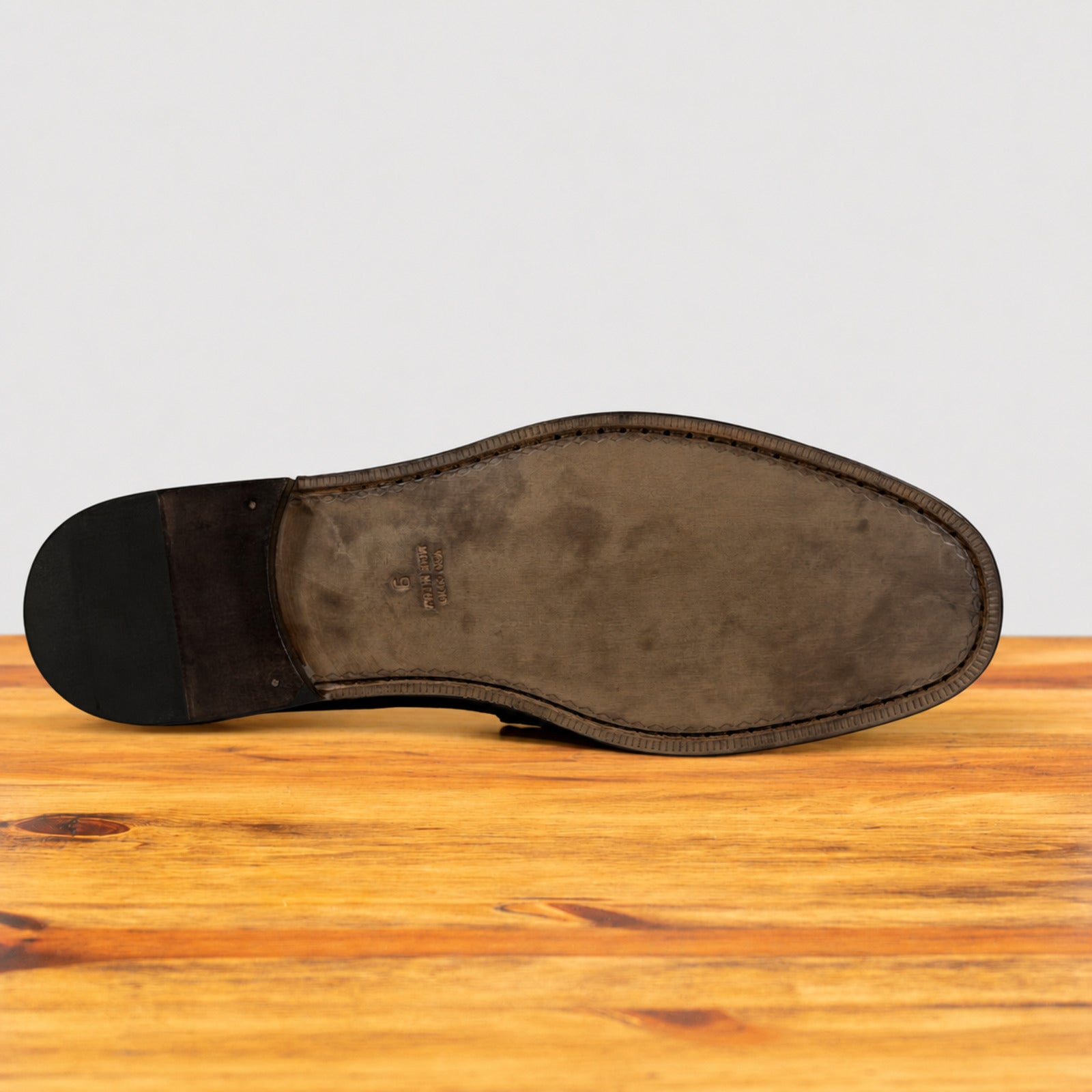 The outsole of a dark brown leather dress shoe is shown from a top-down view, resting on a wooden surface against a light gray background.
