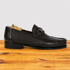 A single dark brown pebbled leather loafer with a horsebit detail on the strap, displayed on a wooden surface against a light gray background.