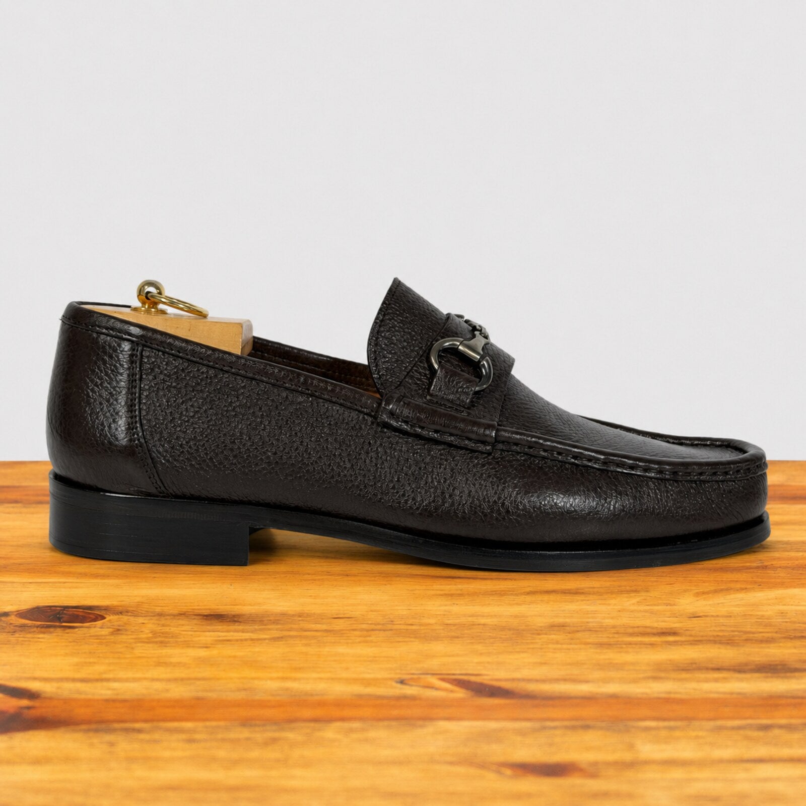 A single dark brown pebbled leather loafer with a horsebit detail on the strap, displayed on a wooden surface against a light gray background.