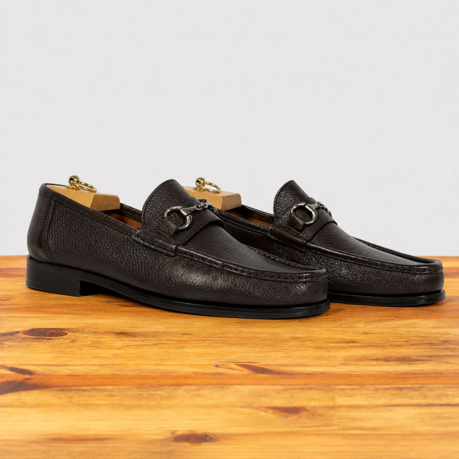 A pair of dark brown pebbled leather penny loafers with gold-tone horsebit detailing, displayed on a wooden surface against a light gray background.
