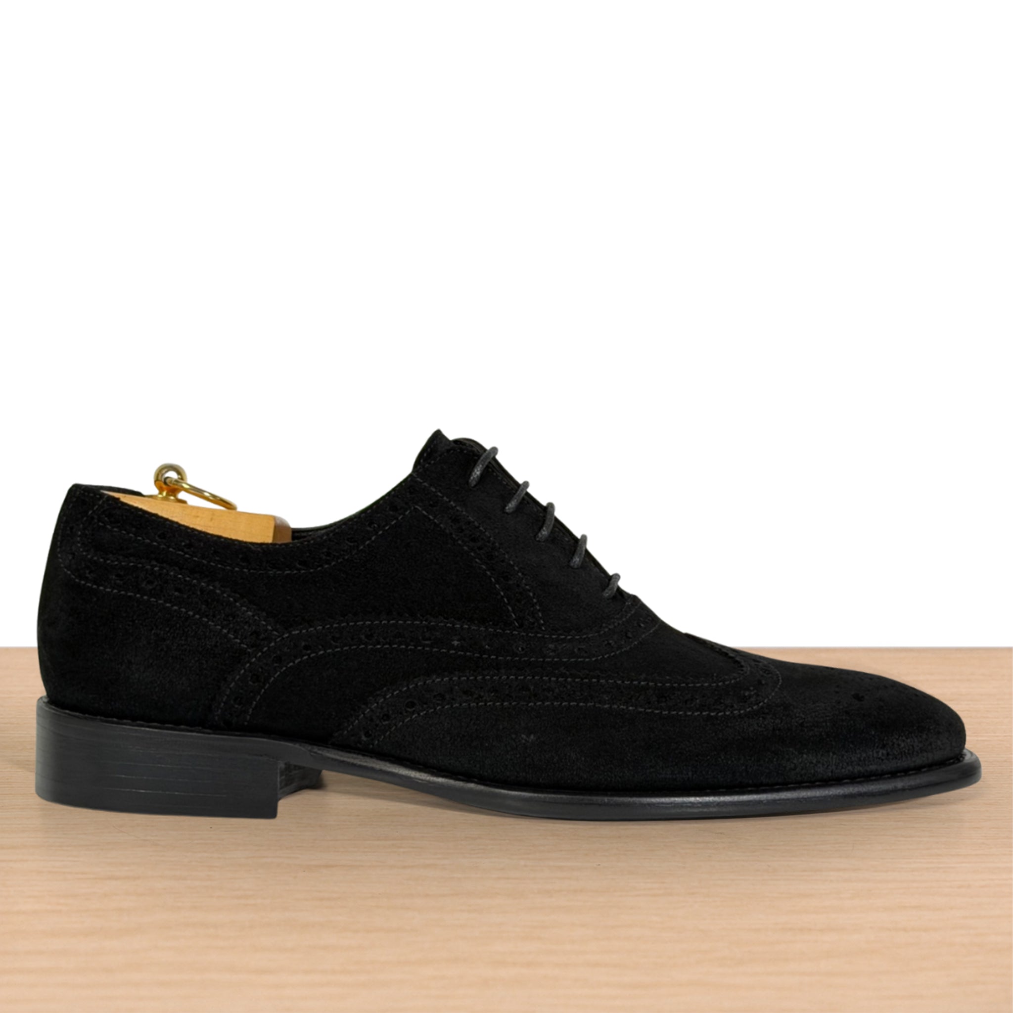 A black suede wingtip oxford shoe with brogue detailing, displayed in profile on a light wood surface against a white background.