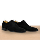 A pair of black suede wingtip oxford shoes with brogue detailing, displayed on a light wood surface against a white background.
