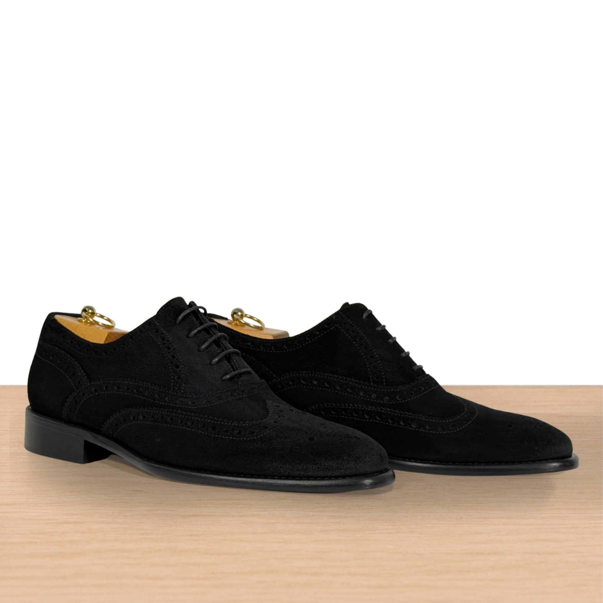 A pair of black suede wingtip oxford shoes with brogue detailing, displayed on a light wood surface against a white background.
