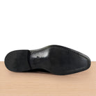 The black leather sole of a dress shoe, showing a square toe and a small embossed logo, resting on a light wood surface against a white background.