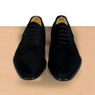 A pair of black suede wingtip oxford shoes with brogue detailing, displayed on wooden shoe trees on a light wood surface against a white background.