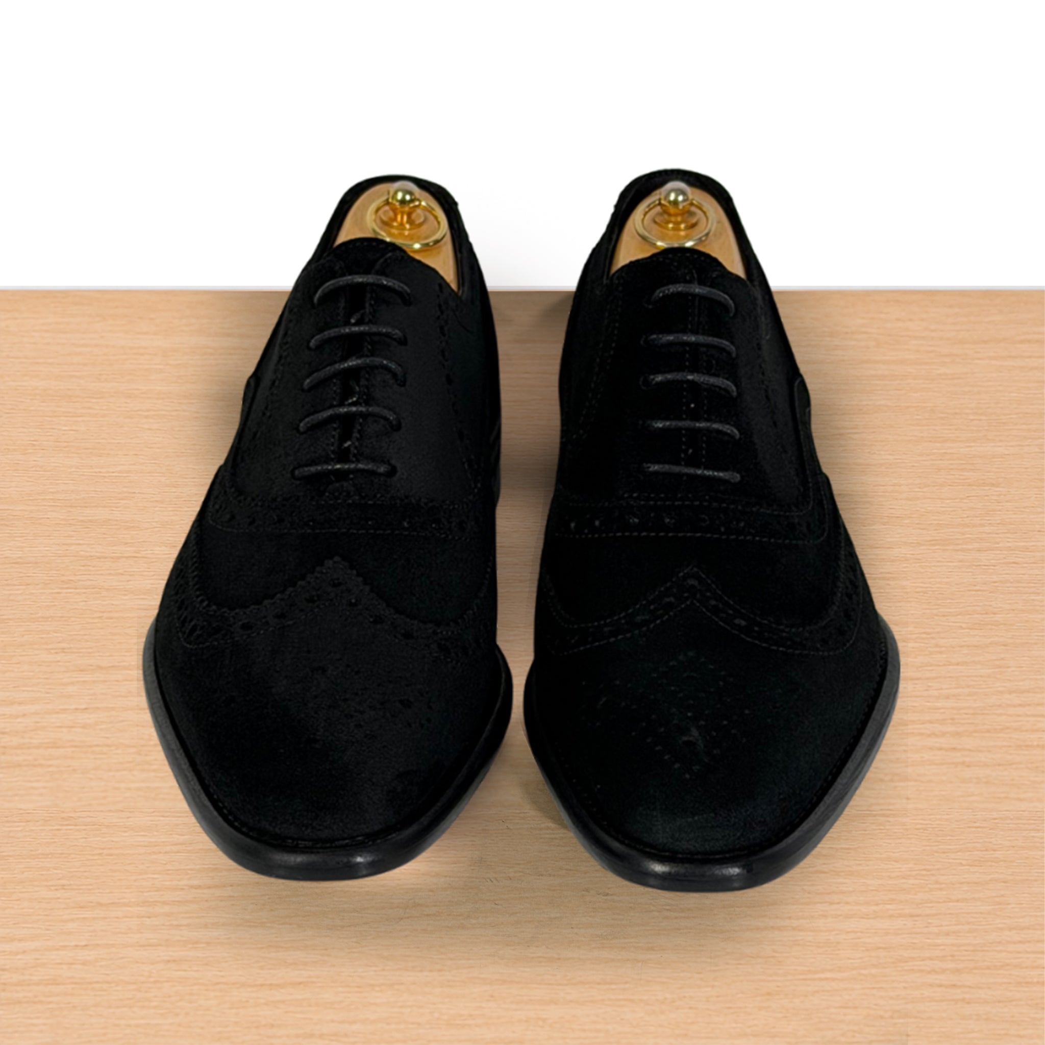 A pair of black suede wingtip oxford shoes with brogue detailing, displayed on wooden shoe trees on a light wood surface against a white background.