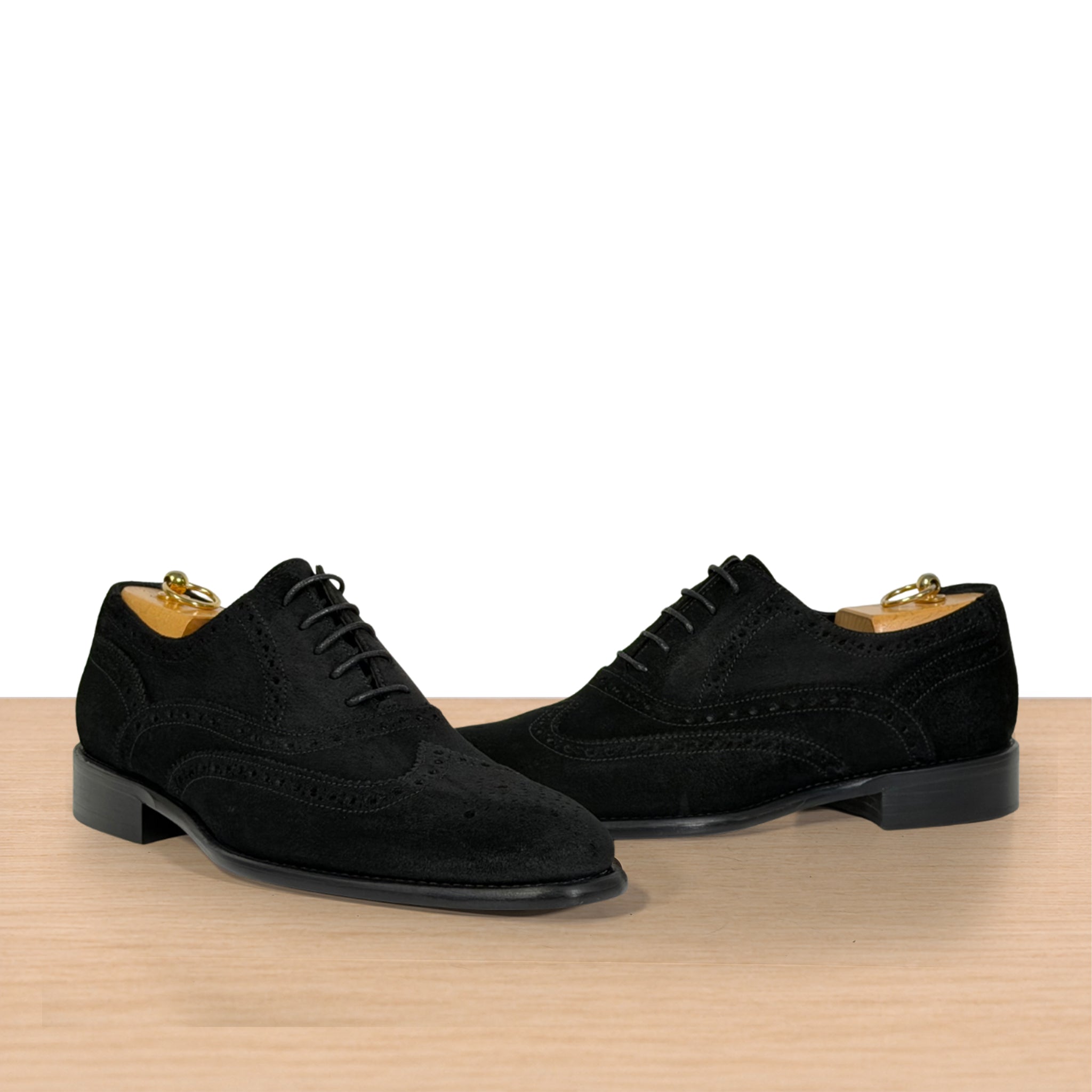 A pair of black suede wingtip oxford shoes displayed on wooden dress forms, set against a clean white background with a light wood surface below.