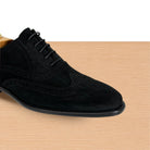 A black suede wingtip oxford shoe displayed on a wooden shoe tree against a light wood-grain surface and white background.