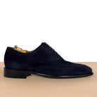 A navy blue suede wingtip oxford shoe with brogue detailing, displayed on a wooden surface against a white background.