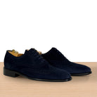 A pair of navy blue suede wingtip oxford shoes with brogue detailing, displayed on a light wood surface against a white background.