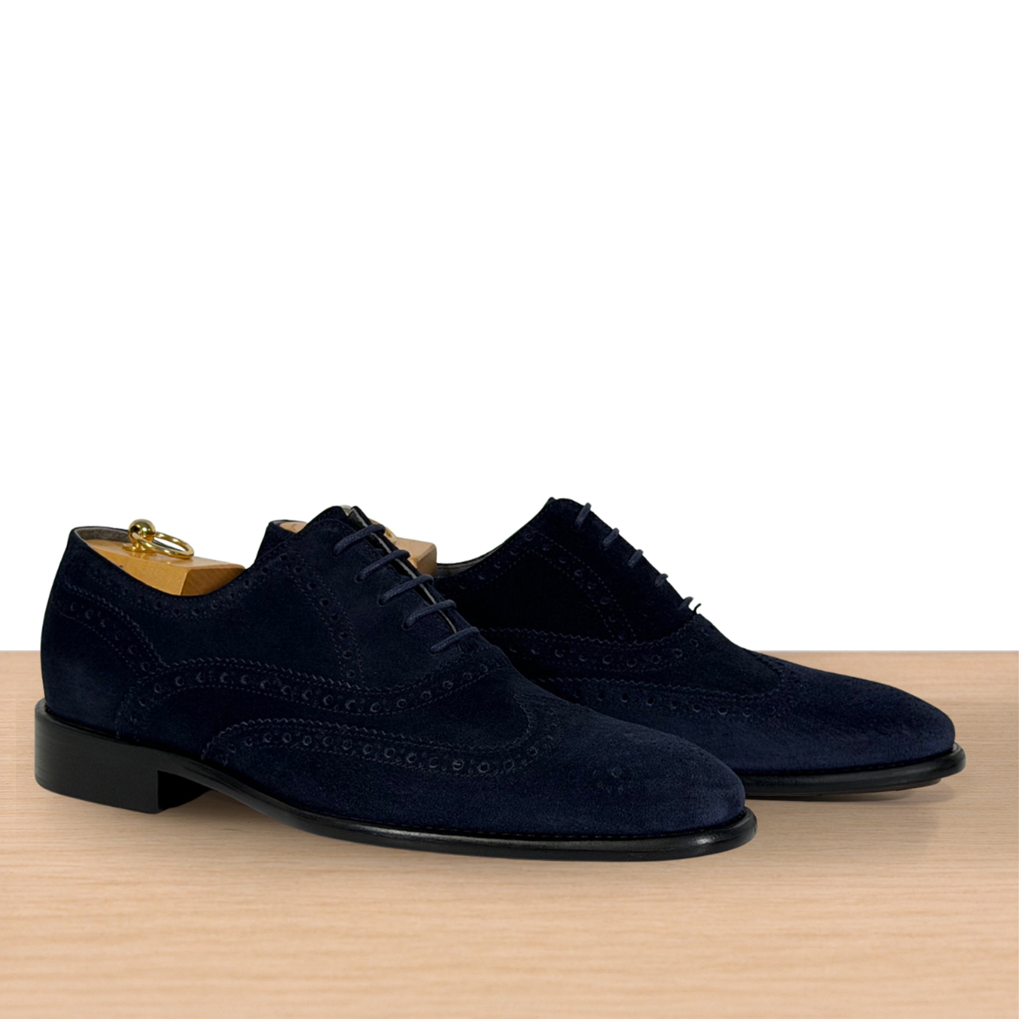 A pair of navy blue suede wingtip oxford shoes with brogue detailing, displayed on a light wood surface against a white background.