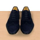 A pair of men's navy blue suede wingtip oxford shoes with black laces and black soles, displayed on a light wood surface against a white background.
