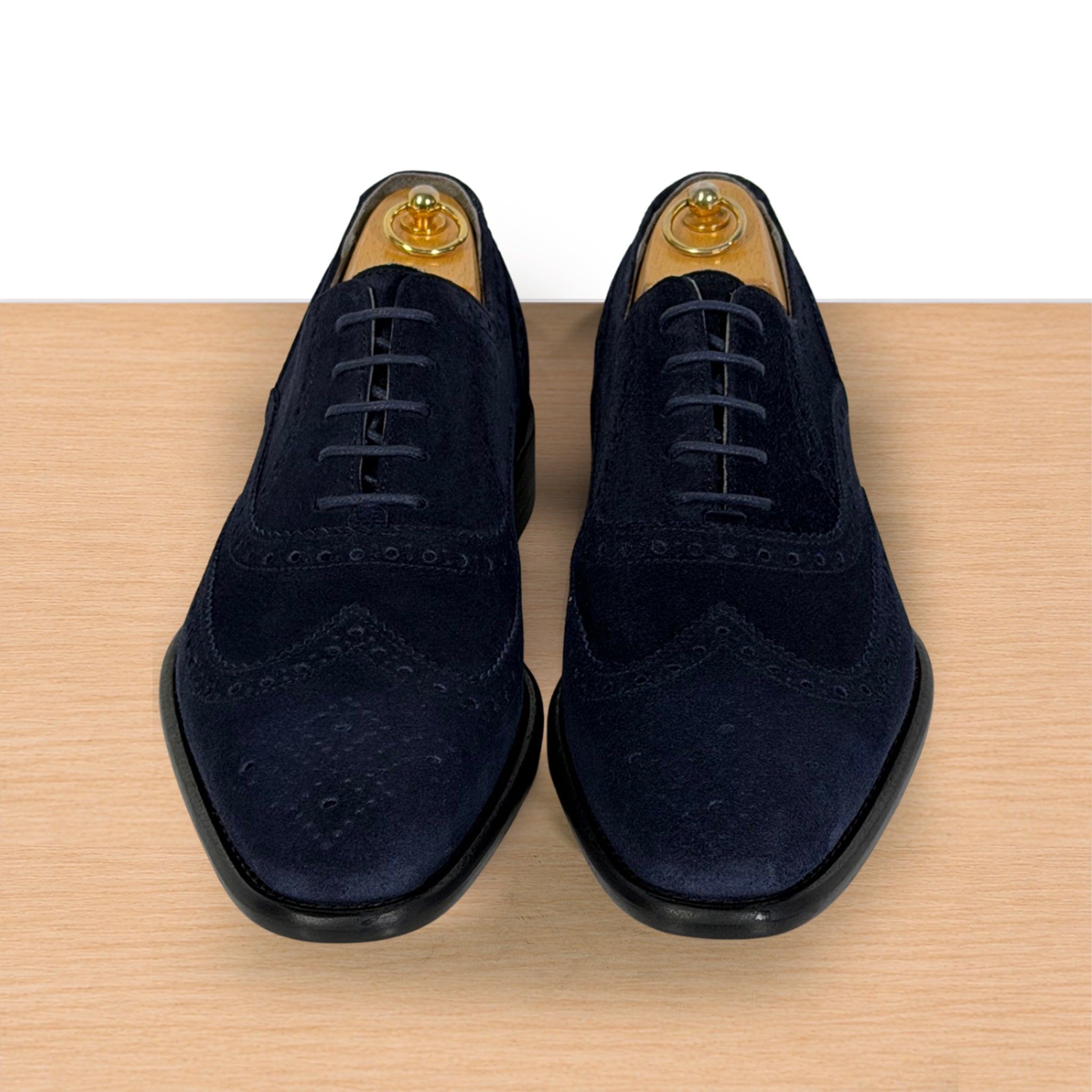 A pair of men's navy blue suede wingtip oxford shoes with black laces and black soles, displayed on a light wood surface against a white background.