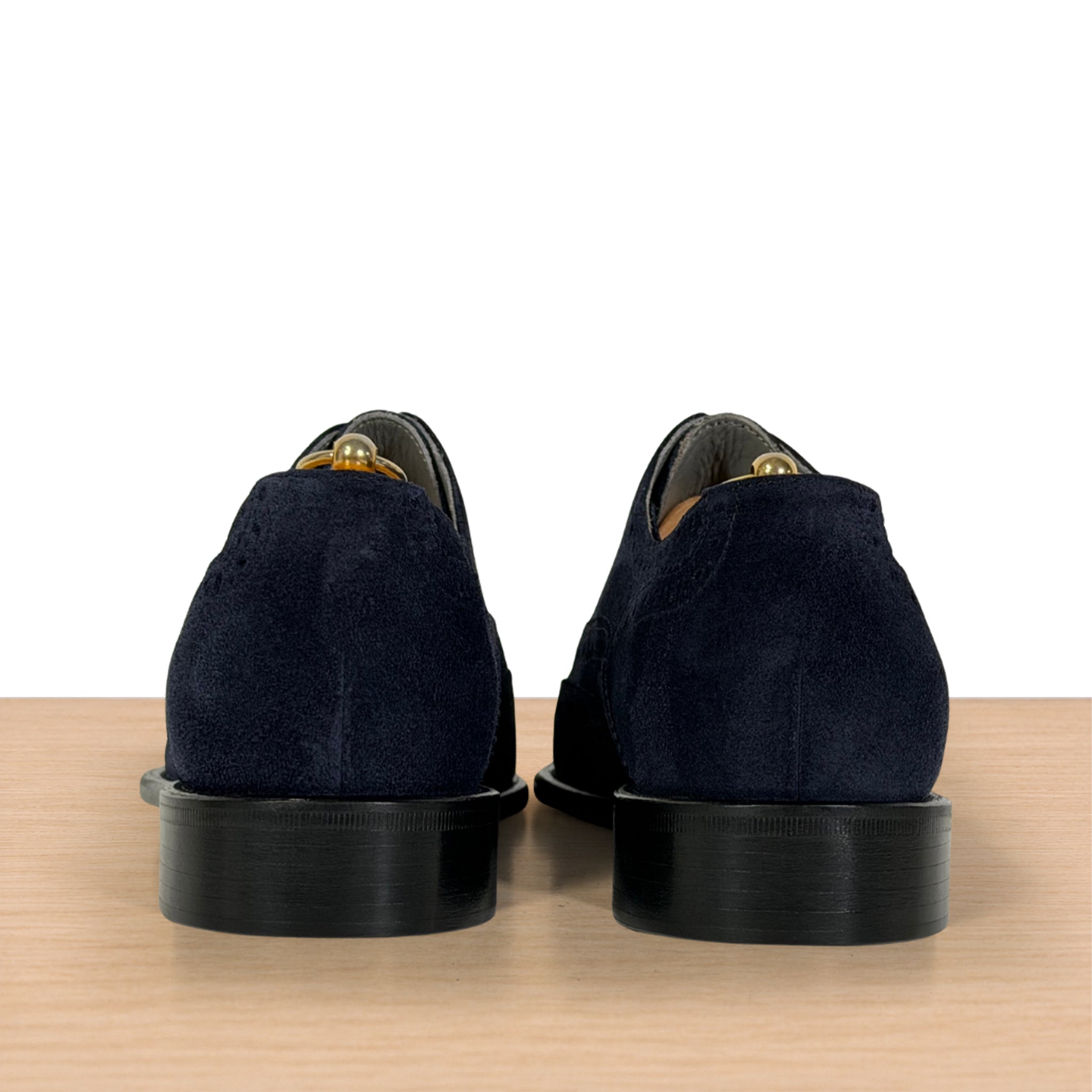 A pair of navy blue suede oxford shoes shown from the back, resting on a light wood surface against a white background.