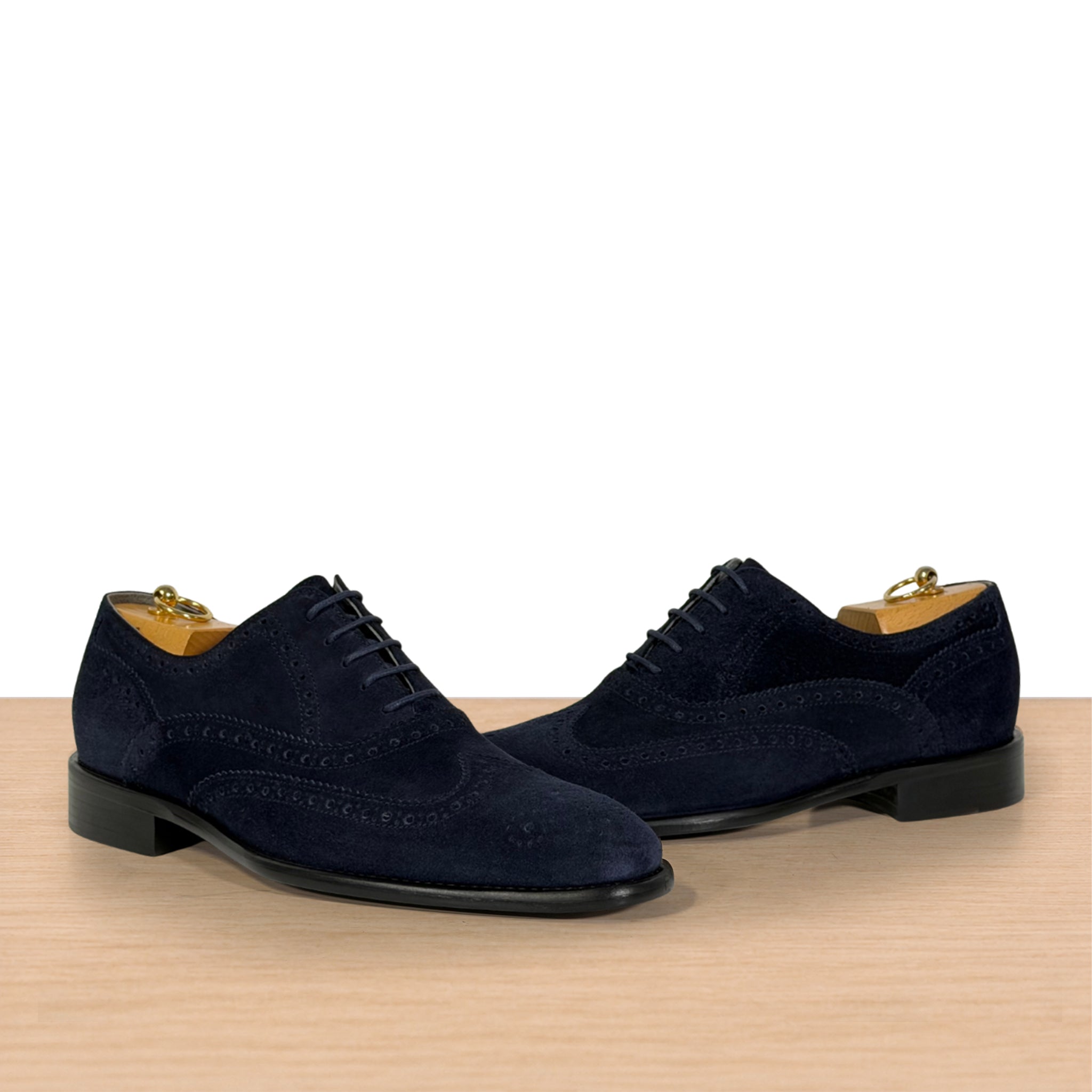 A pair of navy blue suede wingtip oxford shoes with brogue detailing, displayed on a light wood surface against a white background.
