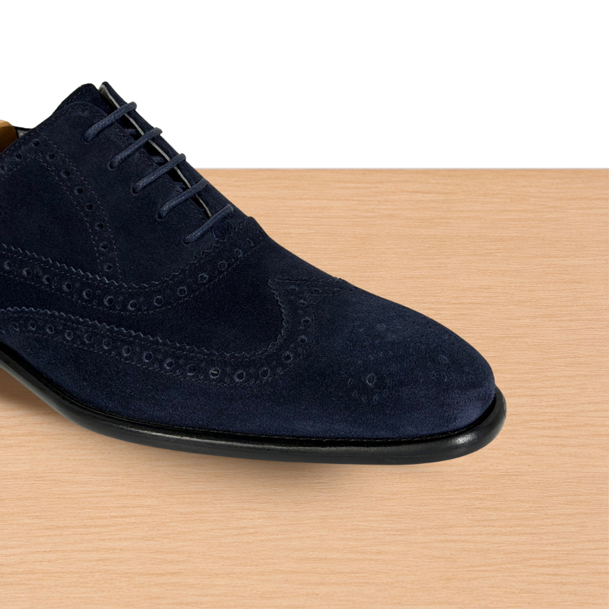 A navy blue suede wingtip oxford shoe with brogue detailing, shown from a side angle on a light wood surface against a white background.